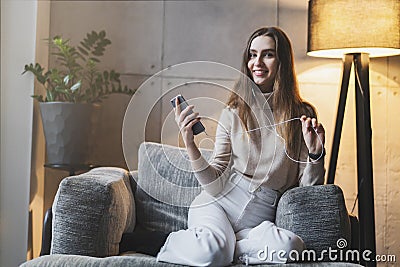 Young female sitting on sofa with smartphone in hand, wearing earphones and listening to music. Girl with mobile phone sending Stock Photo