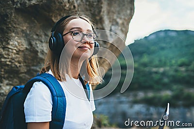 Young female student looks up in hipster glasses and wireless headphones enjoys nature in the mountains while Hiking walk, freedom Stock Photo