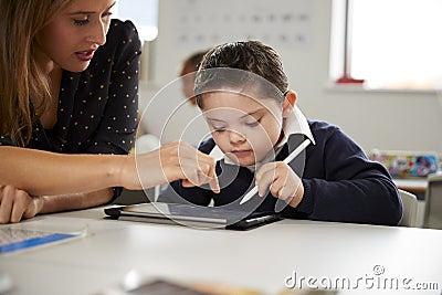 Young female teacher working with a Down syndrome schoolboy sitting at desk using a tablet computer in a primary school classroom, Stock Photo