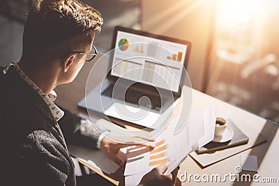 Young finance market analyst in eyeglasses working at sunny office on laptop while sitting at wooden table. Businessman Stock Photo