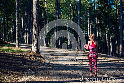 Young fit athletic woman in a forest wearing smart watch and holding yoga mat, walking away from the camera. Stock Photo