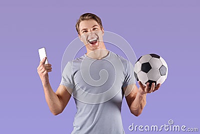 Young football fan watching championship online on smartphone, happy over victory of his team on lilac background Stock Photo