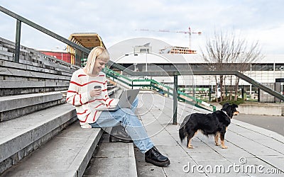 Young freelance expat woman sitting on the stairs of office building drinking coffee and working on laptop computer with her dog Stock Photo