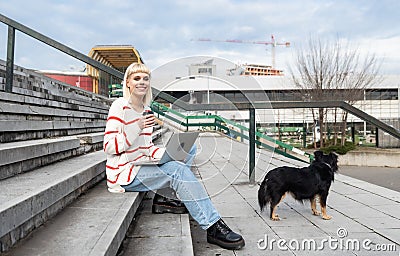 Young freelance expat woman sitting on the stairs of office building drinking coffee and working on laptop computer with her dog Stock Photo