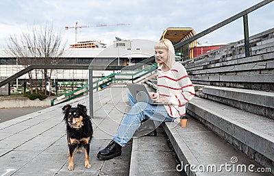 Young freelance expat woman sitting on the stairs of office building drinking coffee and working on laptop computer with her dog Stock Photo