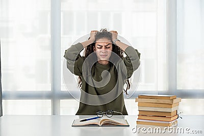 Young frustrated school girl sitting at desk with pile of books feeling sad that she need to read. Female first year college Stock Photo