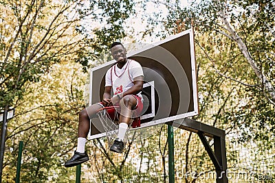 Young funny African American basketball player in a white T-shirt and red shorts sits in a basket on the basketball Stock Photo