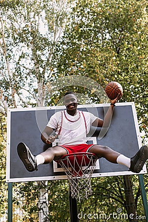 Young funny African American basketball player in a white T-shirt and red shorts sits in a basket on the basketball Stock Photo