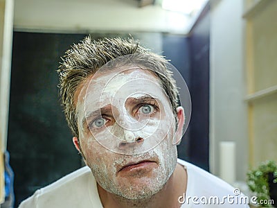 Young funny attractive man at home bathroom looking at toilet mirror applying facemask in his face finding himself weird and ugly Stock Photo