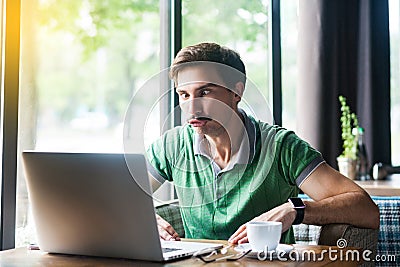 Young funny businessman in green t-shirt sitting and looking at laptop display with stupid crazy face and crossed eyes Stock Photo