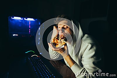 Young gamer sits at home at the table at the computer and eats pizza. Young man eating pizza at break after computer game. Night Stock Photo