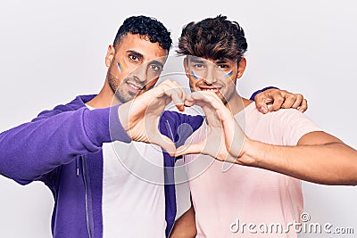 Young gay couple drawing rainbow lgtbq flag on face smiling in love doing heart symbol shape with hands Stock Photo