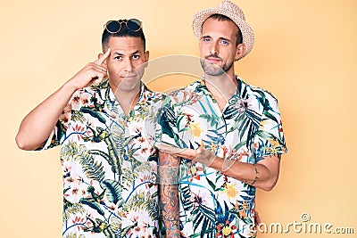 Young gay couple of two men wearing summer hat and hawaiian shirt confused and annoyed with open palm showing copy space and Stock Photo