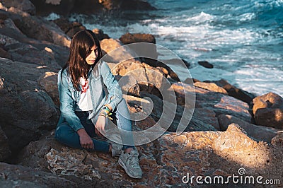 Young girl alone sitting on rocks by the sea in relaxed pose Stock Photo