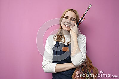 Young girl artist holds brushes and a palette and smiles on a pink background, student of art school, profession of an artist Stock Photo