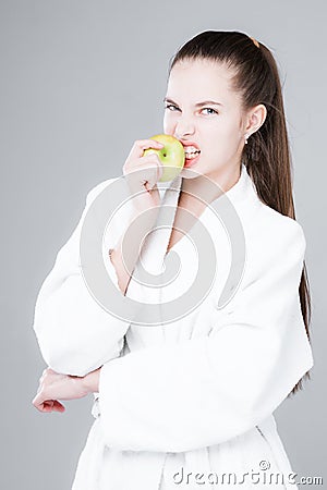 A young girl bites an apple, stands in a white robe on a gray background. Dental health and proper nutrition concept Stock Photo