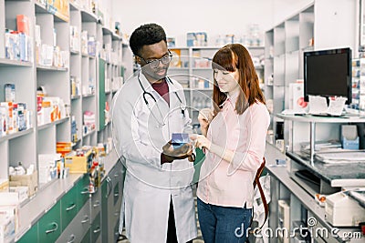 Young girl client who is satisfied of recommended medicines in pharmacy, while standing together with African man Stock Photo