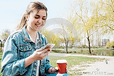 A young girl in a denim jacket walks down the street with coffee and a phone in her hands. Girl in glasses and a denim jacket on Stock Photo