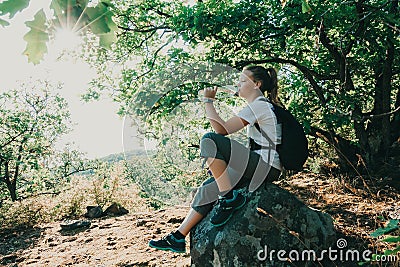 Girl drinking mineral water sitting on stone in forest. Hiker resting with aqua bottle during hike. Thirst quench beverage Stock Photo