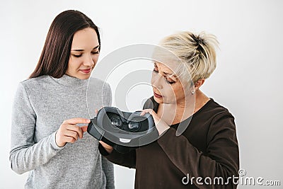 A young girl explains to an elderly woman how to use virtual reality glasses. The older generation and new technologies. Stock Photo