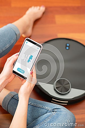 Young girl feet at home using a smart phone to control a round smart automatic vacuum, machine housework robot cleaning the floor Stock Photo