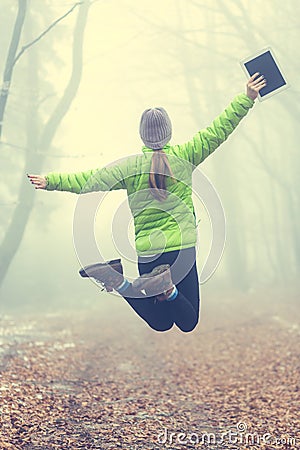 Young girl jumps up with her hands up - Active modern lifestyle - tablet in hand - magic misty forest in autumn mountains. Stock Photo