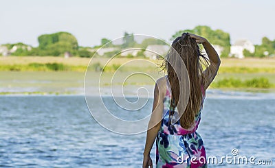 A young girl with long hair turned her back and stares into the distance on the river bank. Stock Photo