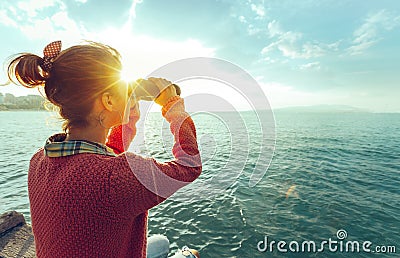 Young Girl Looking Through Binoculars At The Sea On A Bright Sunny Day, Rear View. Wanderlust Travel Journey Concept Stock Photo
