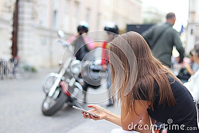 A young girl is looking into a smartphone that is holding. A woman sits on a bench in the center of an ancient city against a moto Stock Photo