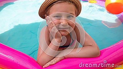 Young girl playing in colorful rainbow inflatable swimming pool.Child in straw hat and pink swimsuit in clear blue water. Stock Photo