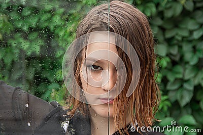 A young girl with predatory look and wet hair is looking at the camera. Wet glass with drops of water divides her face in half. Stock Photo