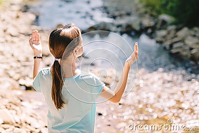 Young girl raises her arms praying on the banks of a mountain stream Stock Photo