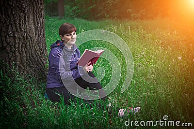 Young girl reading a book sitting on the grass near the oak at sunset. Summer vacation and camping. Fabulous beautiful composition Stock Photo