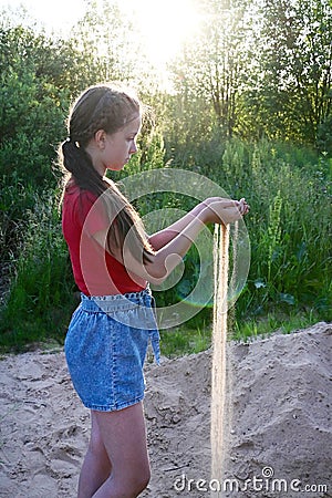 young girl in red T-shirt and denim shorts holds sand in her palms. The sand sifts through my fingers Stock Photo