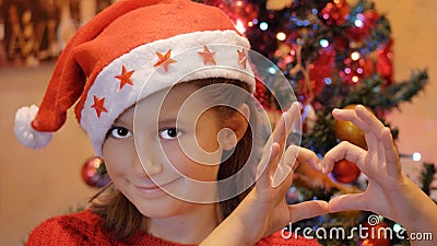 Young girl with Sants Claus Hat doing heart shape with hands with christmas tree as background Stock Photo