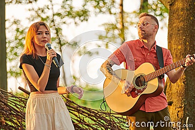 Young girl singer holds microphone in hand with man guitarist plays rock music at outdoor festival Editorial Stock Photo