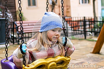 A young girl sits on a swing in a playground, her expression is thoughtful and introspective. Stock Photo