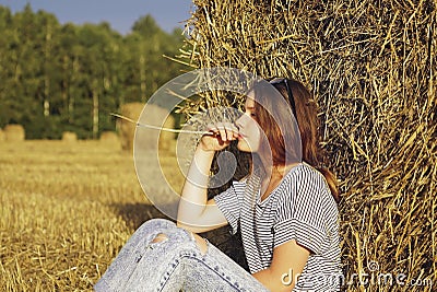 A young girl is sitting near a round stack of straw in a mown field of rye Stock Photo