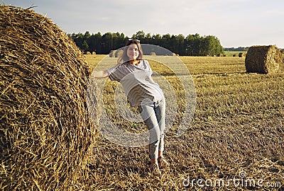 A young girl stands near a round stack of straw on a mown field of rye Stock Photo