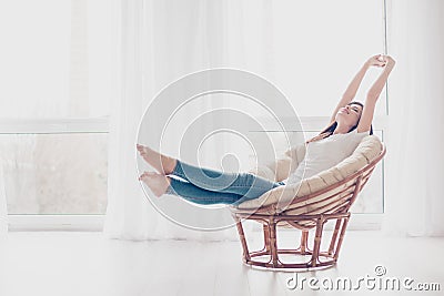 Young girl is stretching in modern armchair in light livingroom. She is sleepy and dreamy, wearing casual clothes Stock Photo