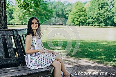 A young girl in a sundress sitting on a park bench. Beautiful woman in a summer white dress and sandals. Girl sitting on a bench Stock Photo