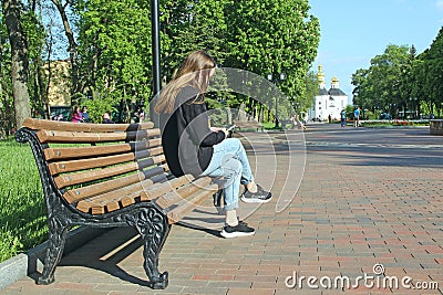 Young girl thoughtfully looking at smartphone sitting on bench in city park Editorial Stock Photo