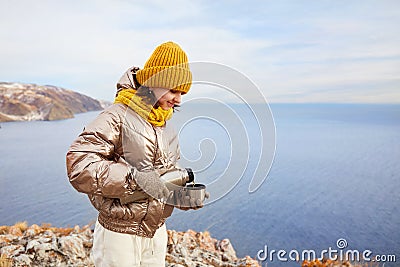 A young girl traveler pours hot tea into a mug from a thermos in nature. Mountain winter landscape. Stock Photo