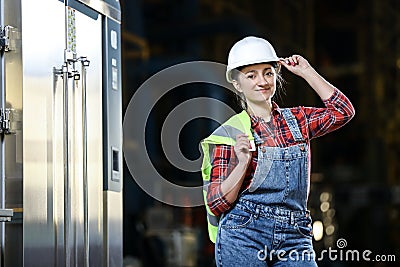 Young girl in a work dress and white hard hat in a factory. Woman in a work uniform holding reflective vest in a storage. Stock Photo