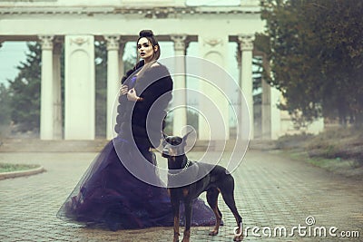 Glam lady wearing luxurious sequin violet evening gown and fur coat standing on the alley in the park with her Doberman pinscher Stock Photo