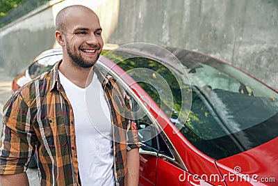 Young good looking bold bearded guy standing outdoors near his red car smiling. wearing yellow shirt Stock Photo