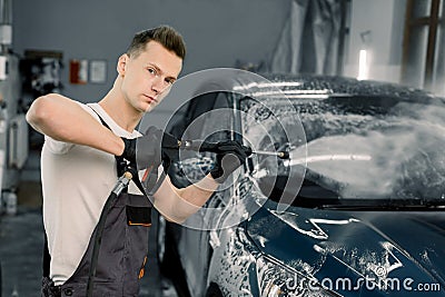 Young good-looking man worker washing luxury dark blue car on a car wash, looking at camera and holding high pressure Stock Photo