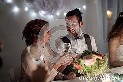Young groom and his mother sitting at a table on a wedding, clinking glasses. Stock Photo