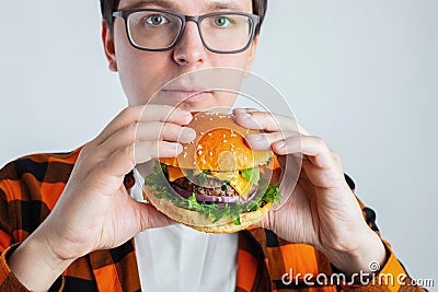 A young guy with glasses holding a fresh Burger. A very hungry student eats fast food. Hot helpful food. The concept of gluttony a Stock Photo