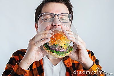 A young guy with glasses holding a fresh Burger. A very hungry student eats fast food. Hot helpful food. The concept of gluttony a Stock Photo